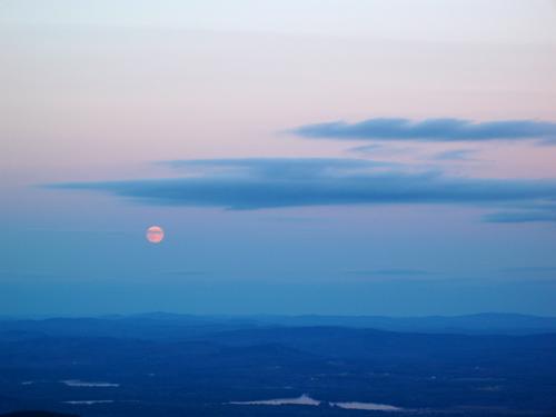 the moon rises amidst soft sunset color as seen from the summit of Kearsarge North in New Hampshire