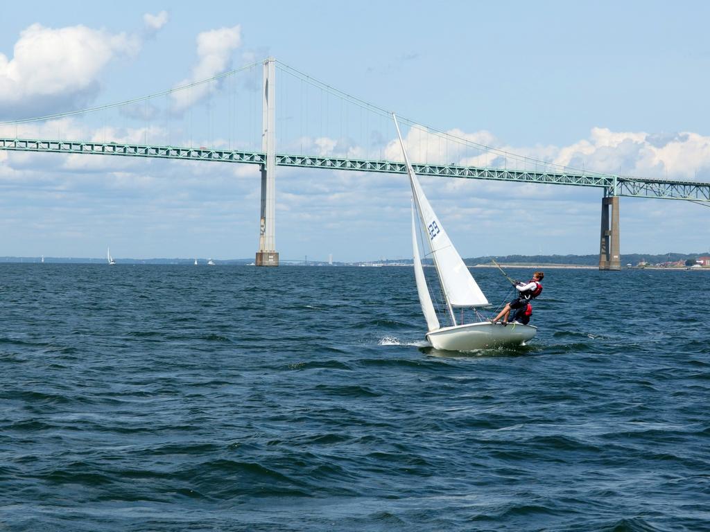 young sailors master a difficult craft, one performing a gymnastic stunt 
to counter the tipping force of the wind, at Narragansett Bay in Rhode Island